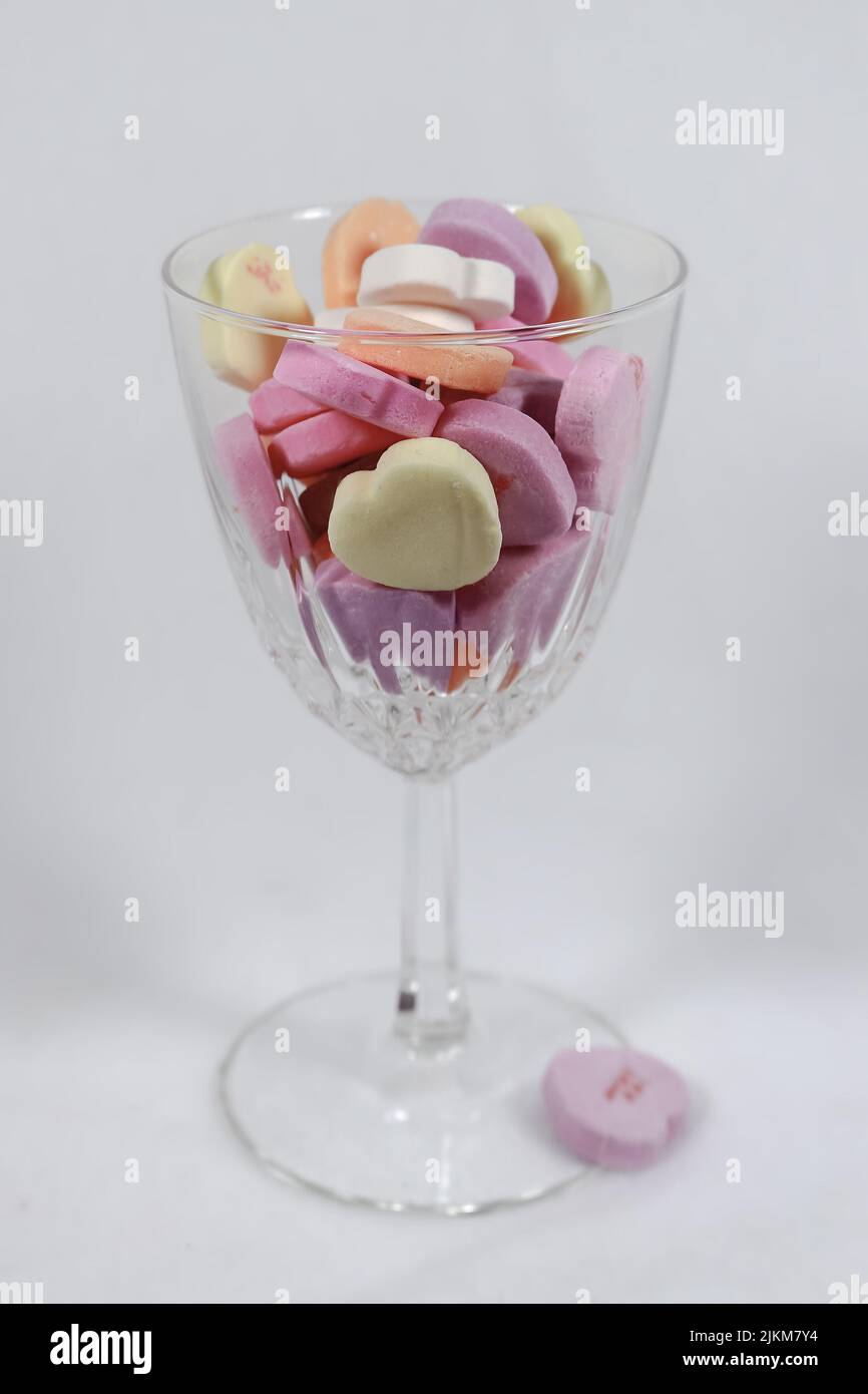 Crystal wine glass holding pastel colored candy hearts against a light background - selective focus Stock Photo