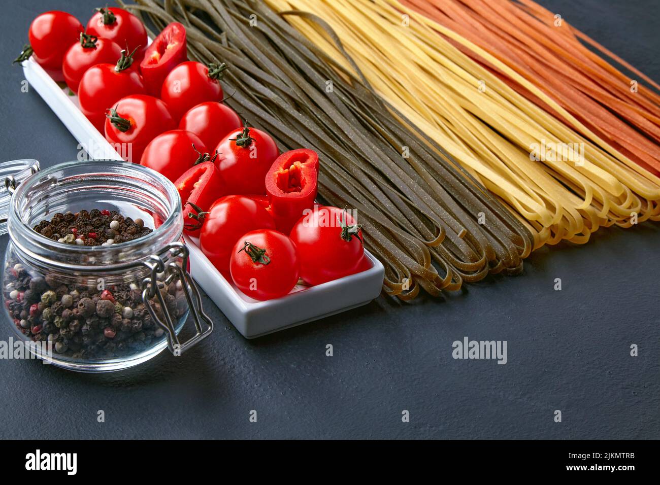 Homemade noodles in different colors from natural vegetable ingredients, an oblong bowl with tomatoes and red paprika and a glass jar with mixed peppe Stock Photo