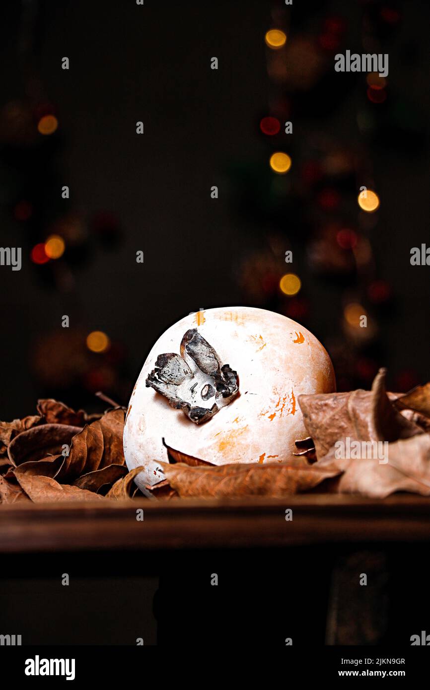 A vertical shot of a persimmon fruit on a table with dried leaves and bokeh lights in the background Stock Photo