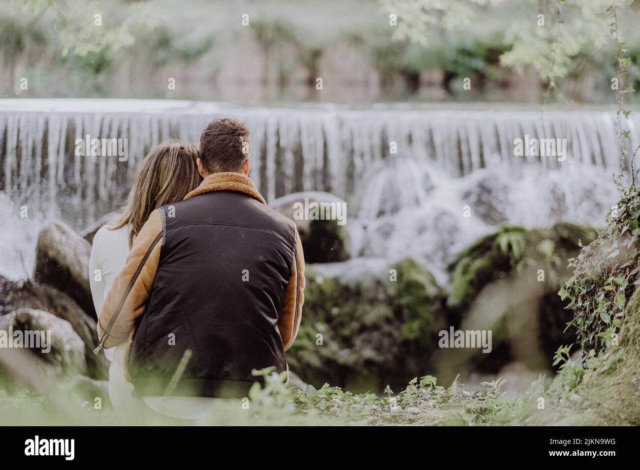 A back view of a couple sitting on the riverside in the park in autumn ...