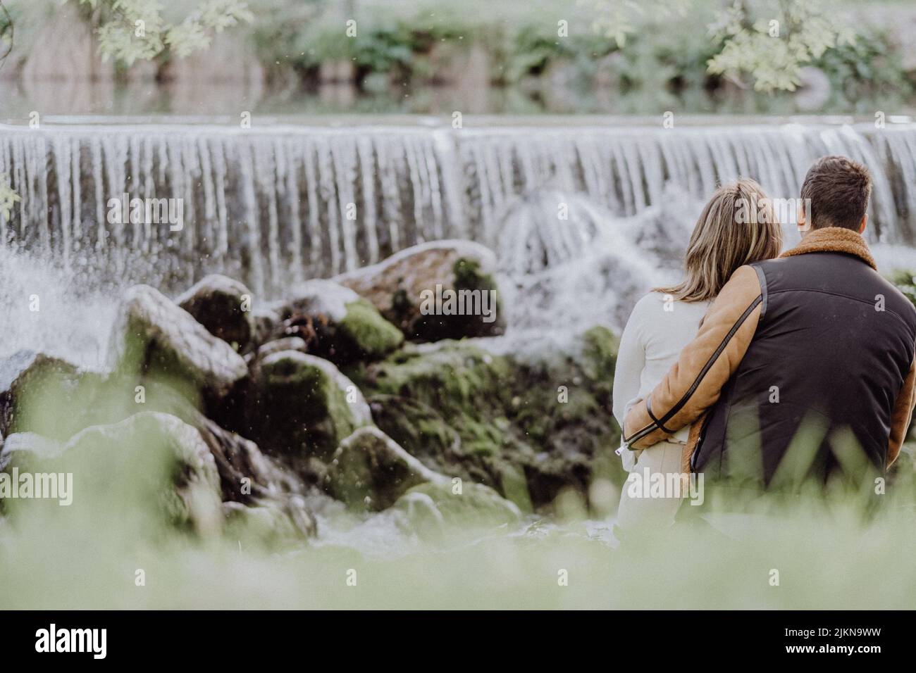 A back view of a couple sitting on the riverside in the park in autumn ...
