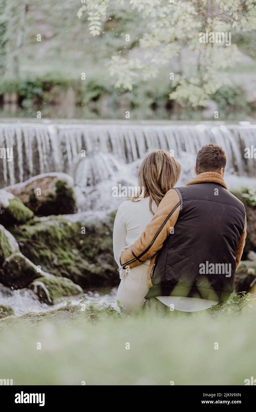 A back view of a couple sitting on the riverside in the park in autumn ...