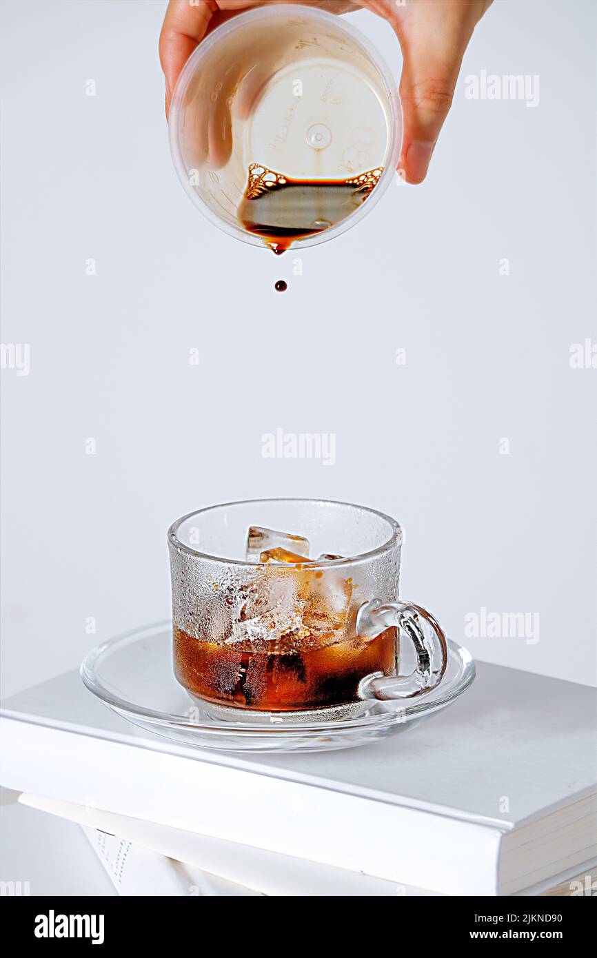 A vertical shot of a hand gradually adding coffee to a cup with ice cubes on a white background Stock Photo