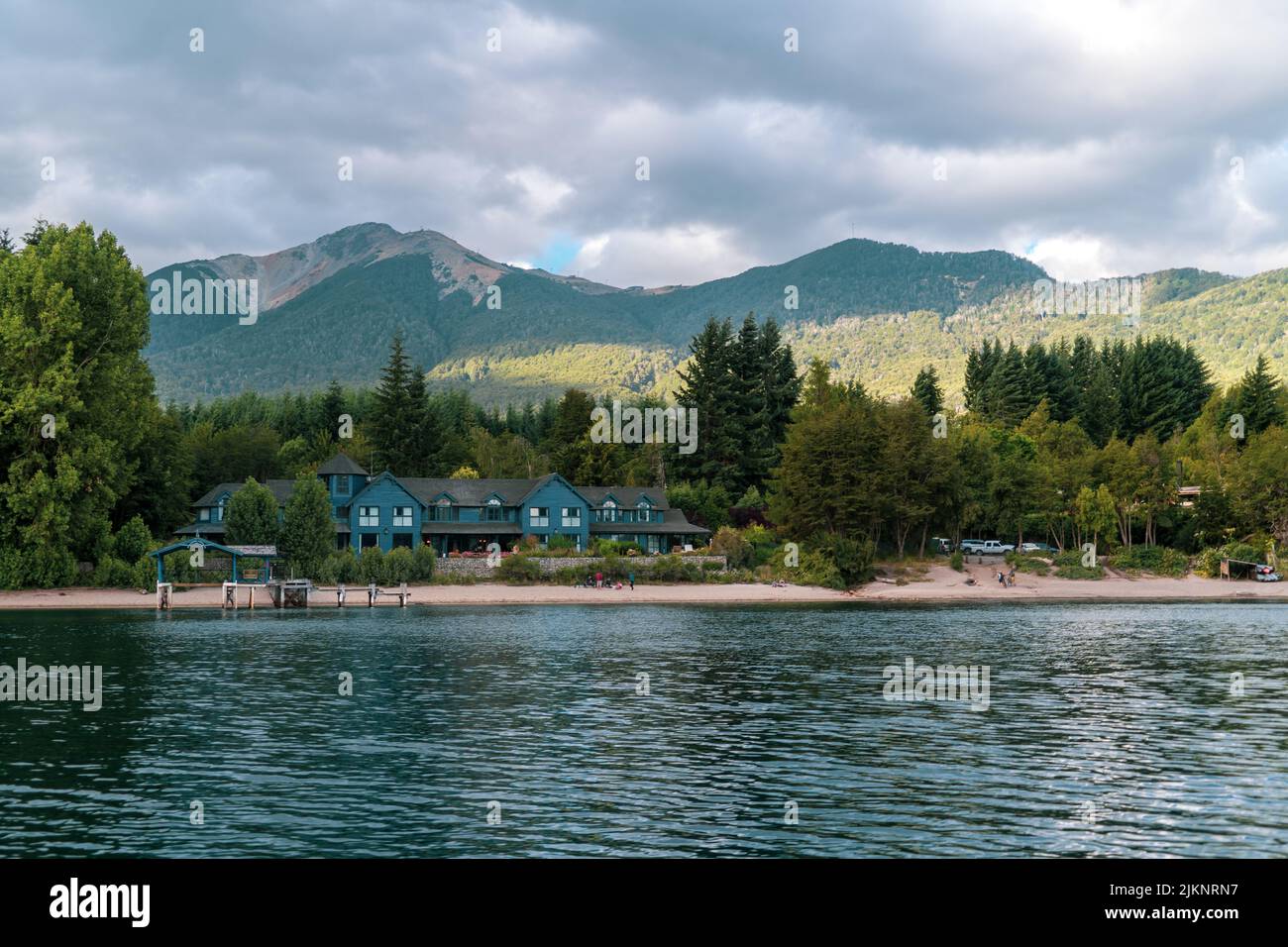The beautiful summer landscape with a hotel on the lakeside surrounded by green trees against mountains. Stock Photo