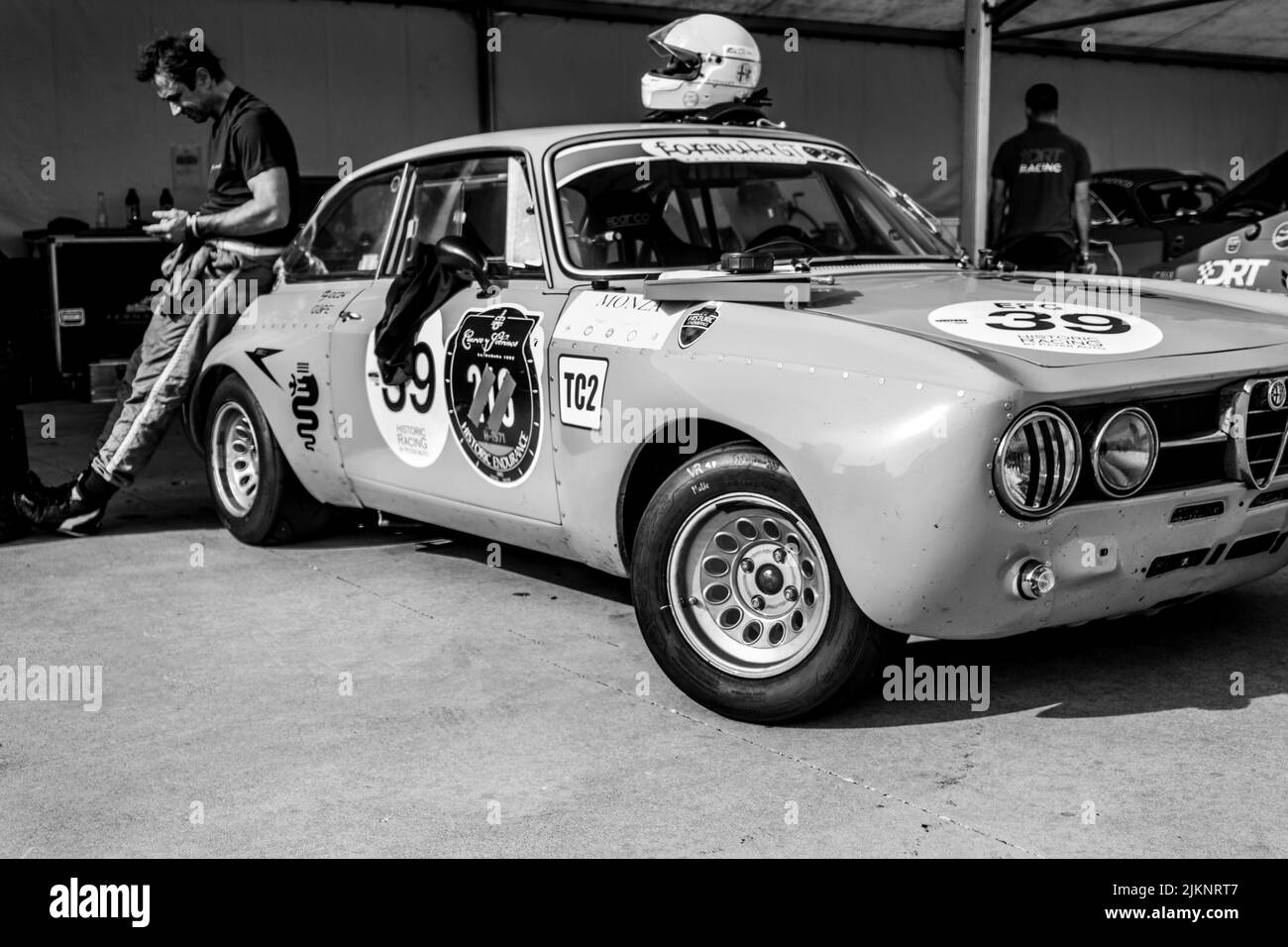 A grayscale shot of a driver relaxing after the race leaning on a vintage car with stickers Stock Photo
