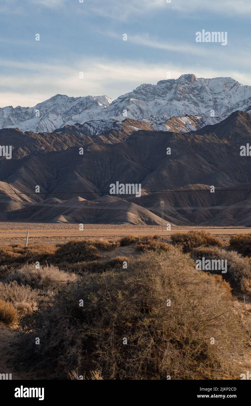 A vertical shot of deserted hills in the background of snowy mountains under the clear cloudless sky Stock Photo