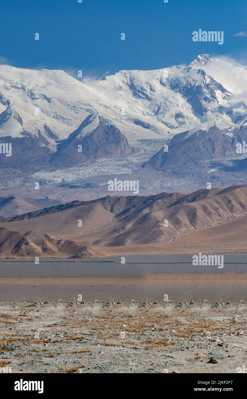 A vertical shot of deserted hills in the background of snowy mountains under the clear cloudless sky Stock Photo