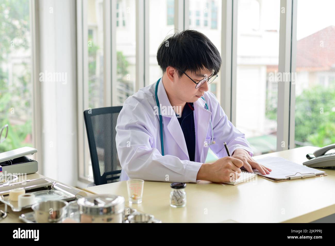 Asian doctor sitting at desk writing recording healthcare treatment on notebook Stock Photo