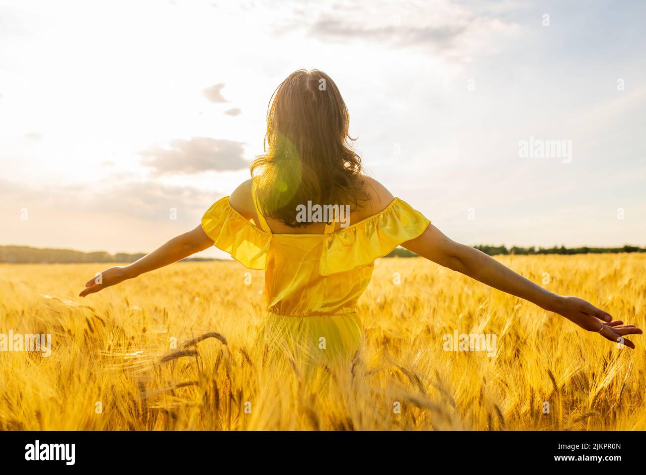 Young woman in yellow dress standing on a wheat field with sunrise on the background Stock Photo