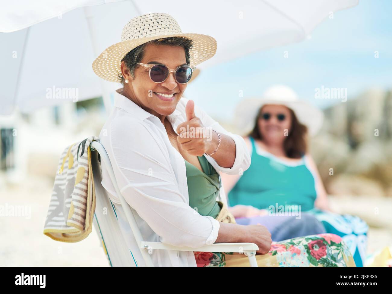 Its a beach day with the girls. a mature woman sitting and making a thumbs up gesture during a day out on the beach with friends. Stock Photo
