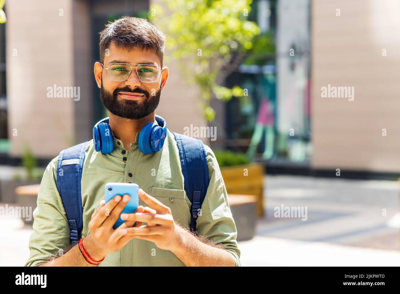 indian student with blue headset and backpack holding smartphone and looking at camera at sunny day Stock Photo