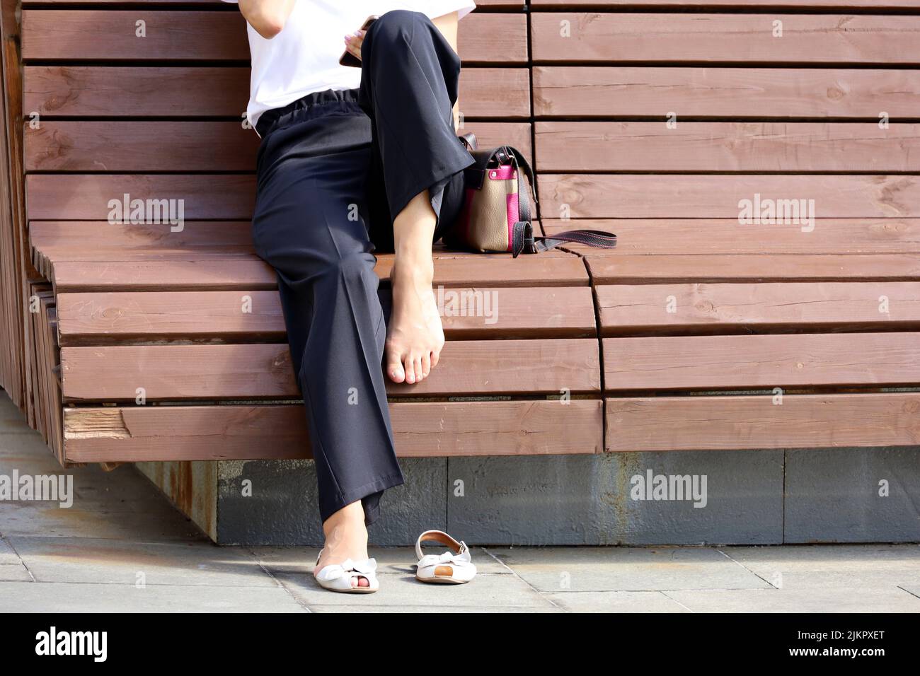 Slim woman with one removed slipper sitting on wooden bench and using smartphone on a street. Tired female legs, summer leisure in city Stock Photo