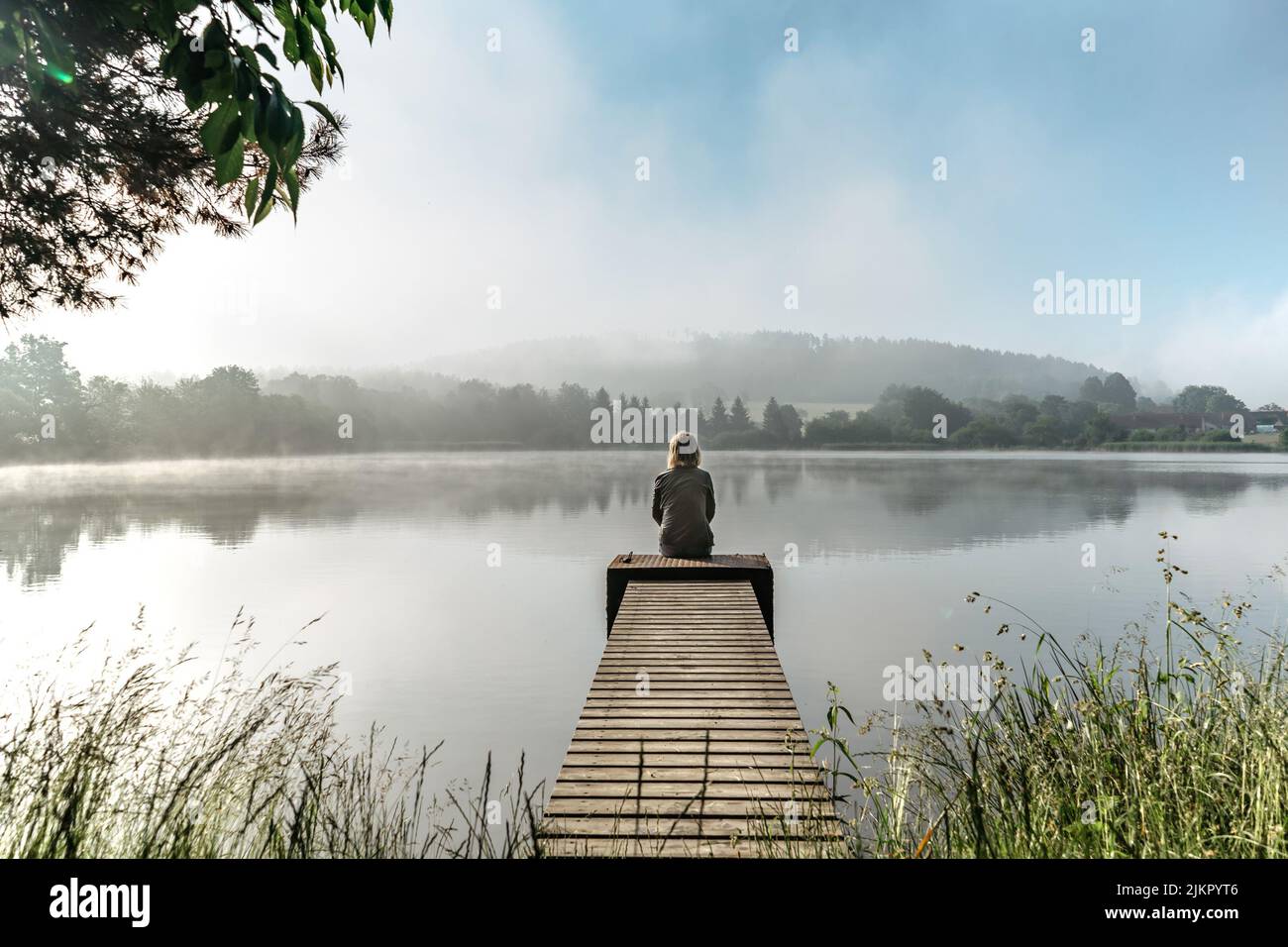 Calm misty morning relaxation by pond.Sitting woman in countryside.Intended female mysterious atmosphere. Spring foggy nature.Silence.Woman feelings Stock Photo