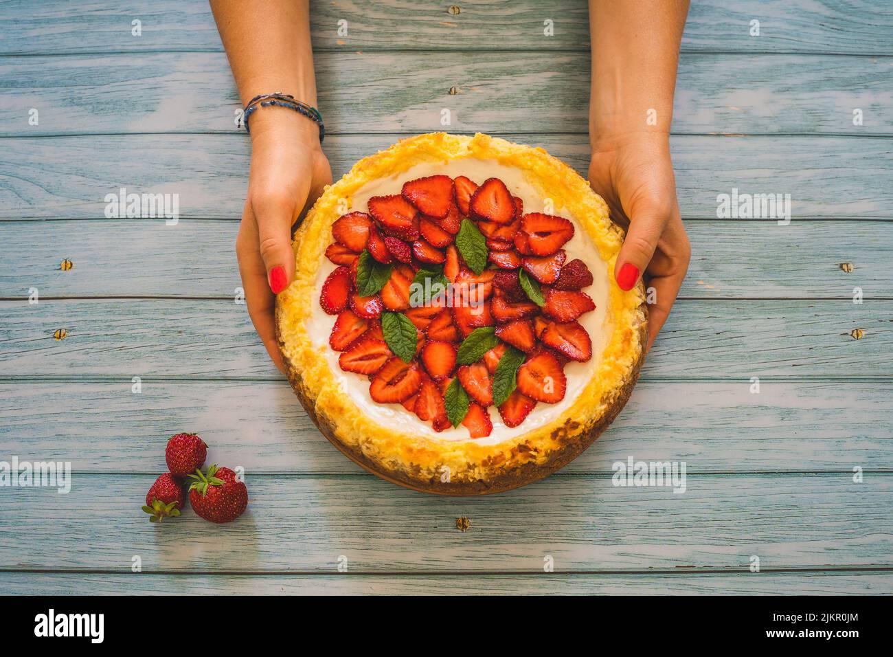 Strawberry homemade cake on wooden table. Woman hand holding delicious cheesecake with strawberries decorated with mint leaves.Top view.Healthy organi Stock Photo