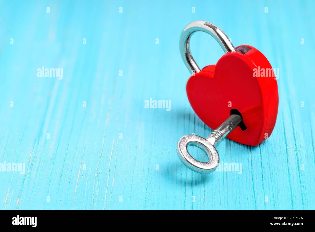 Silver-toned skeleton key in a small red heart-shaped padlock on a blue wooden background with copy space. Symbolic romantic concept. Stock Photo