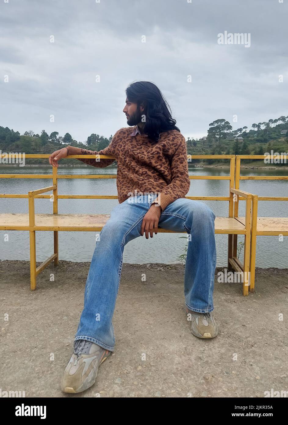 Full length of a long haired and bearded young man looking sideways while sitting on bench next to the lake Stock Photo