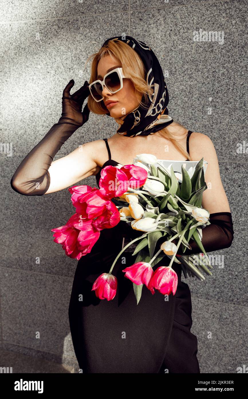 Blonde attractive girl in stylish vintage headscarf, black gloves and dress, lower sunglasses by hand and holding bouquet of fresh pink tulips. Paris Stock Photo