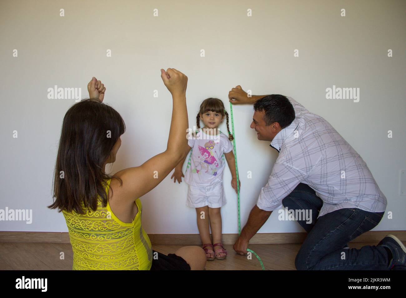 Photo of a dad who measures the height of his daughter with a tape measure while the mother rejoices at the progress achieved. Image of a happy family Stock Photo
