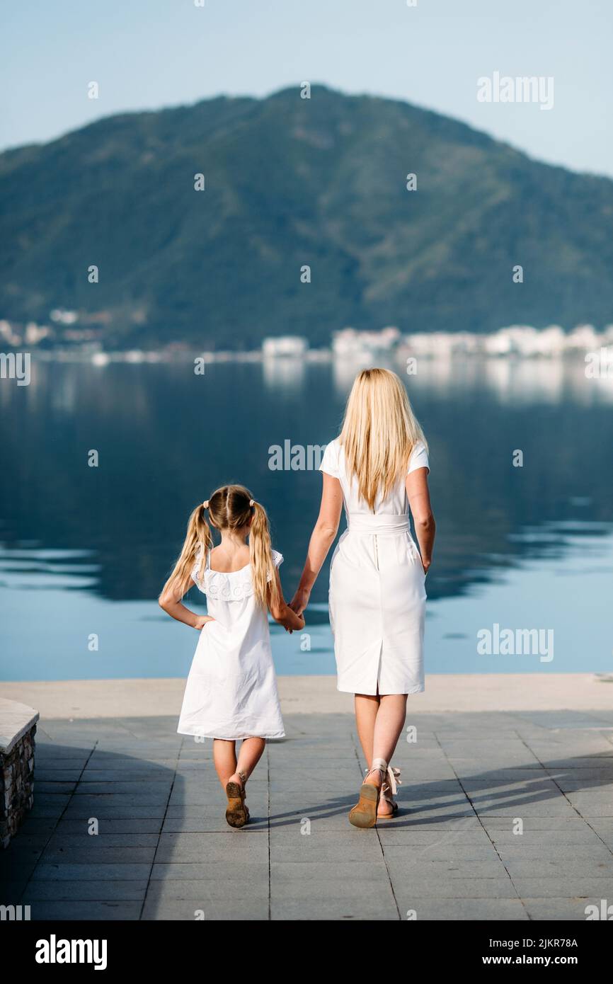 Happy millenial mother and blonde teenager daughter in white family look dresses walking by the harbor and seashore  in a touristic  luxury sea resort Stock Photo