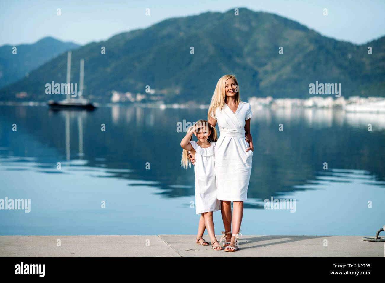 Happy millenial mother and blonde teenager daughter in white family look dresses walking by the harbor and seashore  in a touristic  luxury sea resort Stock Photo