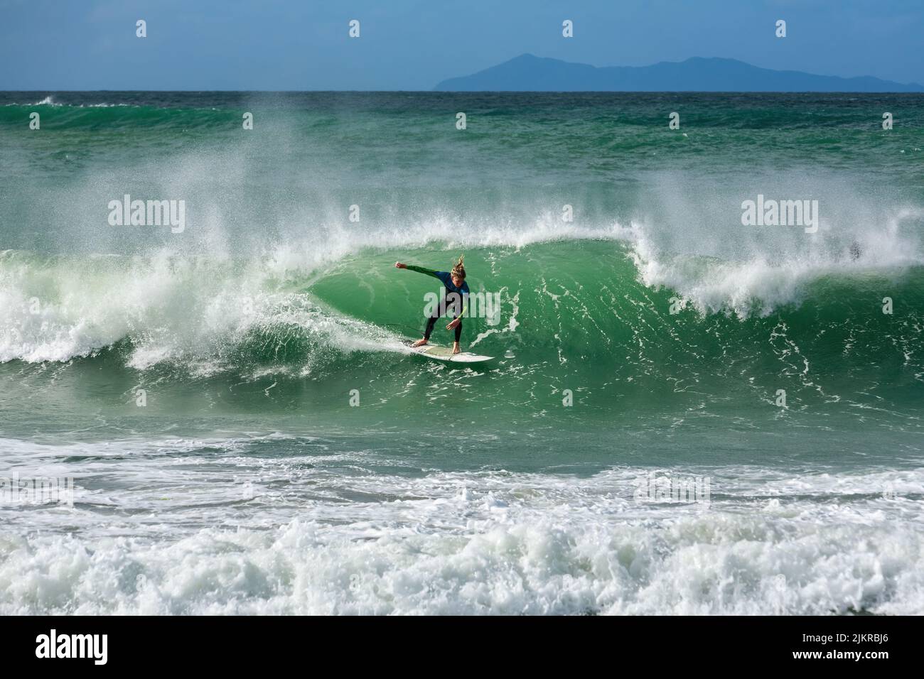 A surfer rides a wave at Mount Maunganui, New Zealand, one of the country's most popular surfing beaches. In the background is Mayor Island Stock Photo