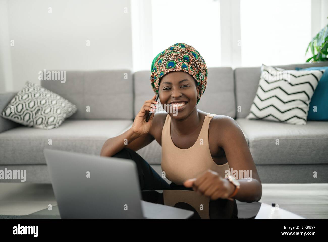 Beautiful young African woman wearing tradition head tie scarf. Sitting at home working on laptop. On phone call smiling and gesturing with hand Stock Photo