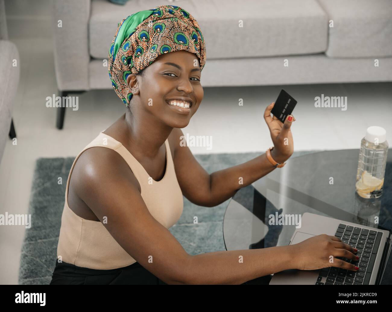 African woman wearing traditional headscarf, making online shopping purchase using laptop and credit card. Sitting by coffee table in lounge at home Stock Photo