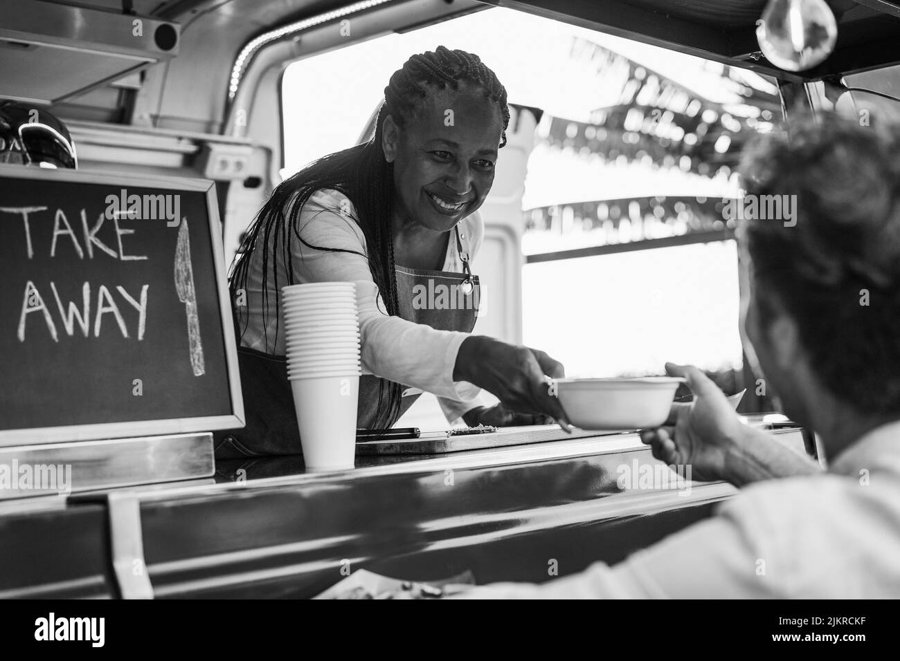 African senior woman serving takeaway food inside food truck - Focus on chef female face - Black and white editing Stock Photo