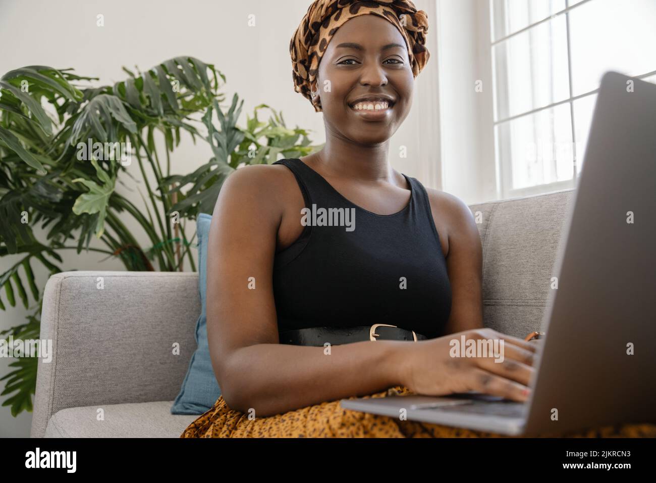 Close up Beautiful traditional young gen z African woman sitting on sofa at home, typing on laptop, smiling, looking at camera Stock Photo