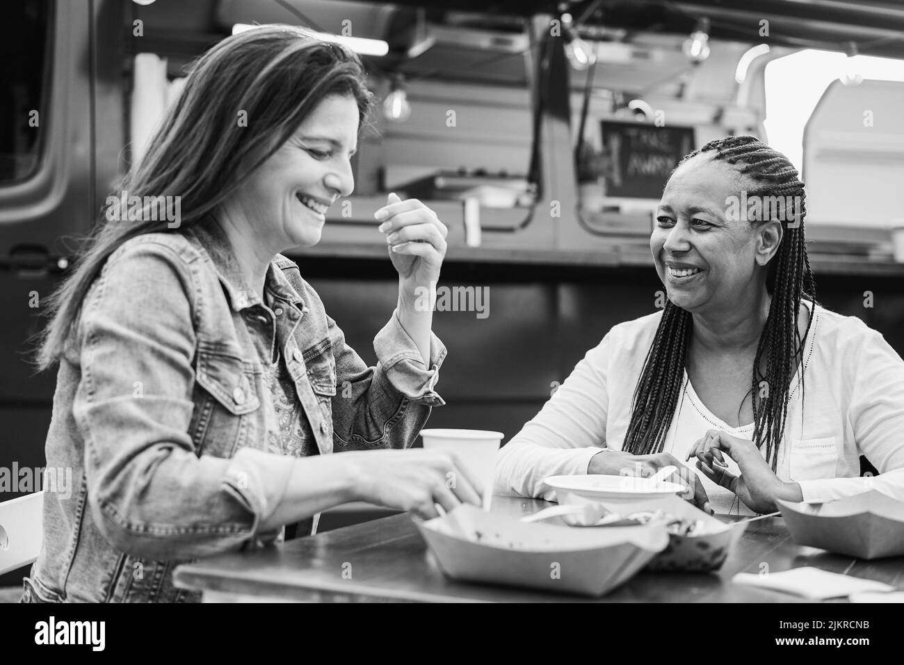 Happy multiracial senior women eating at food truck restaurant outdoor - Focus on african female face - Black and white editing Stock Photo