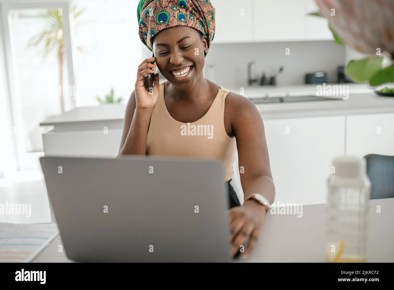 Beautiful young African woman wearing tradition headscarf. Sitting at home working on laptop. On phone call smiling and gesturing with hand Stock Photo