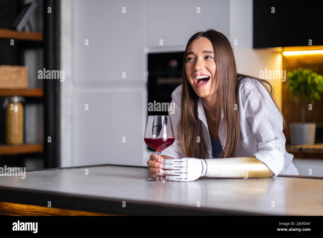 Expressive exited Laughing girl prosthetic arm with glass of wine at home, woman leads normal life artificial prosthetic limb, people with special Stock Photo