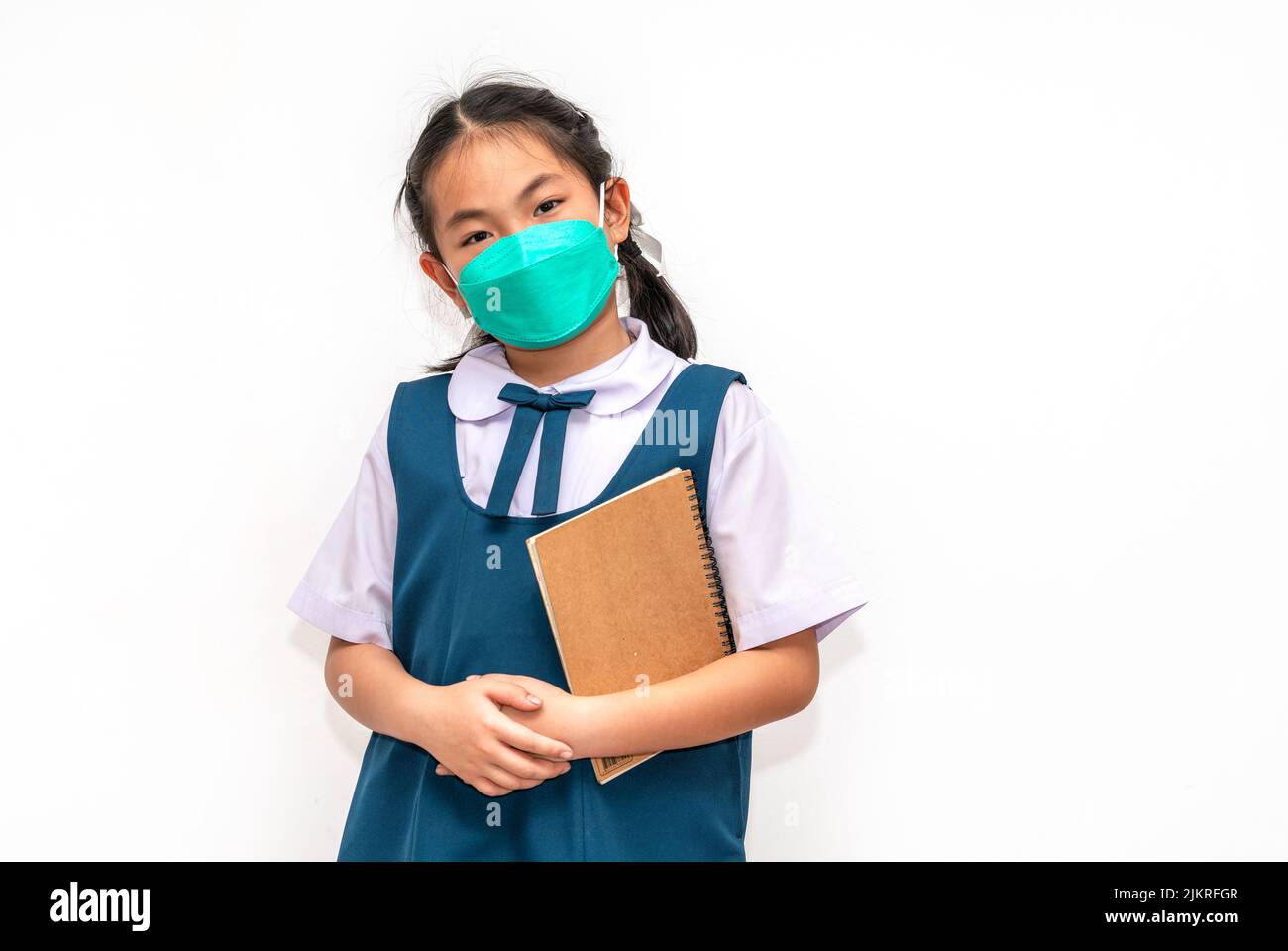 Portrait student Asian child girl in uniform, holding a book, wearing face mask for protection in era of covid virus, isolated student child girl on w Stock Photo