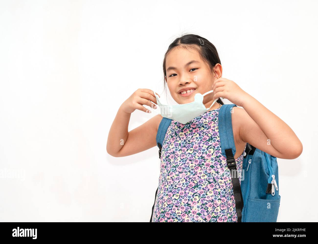 Portrait Asian child girl is holding and willing to wear a facemask, carrying a backpack to go to school, isolated portrait image on white background. Stock Photo