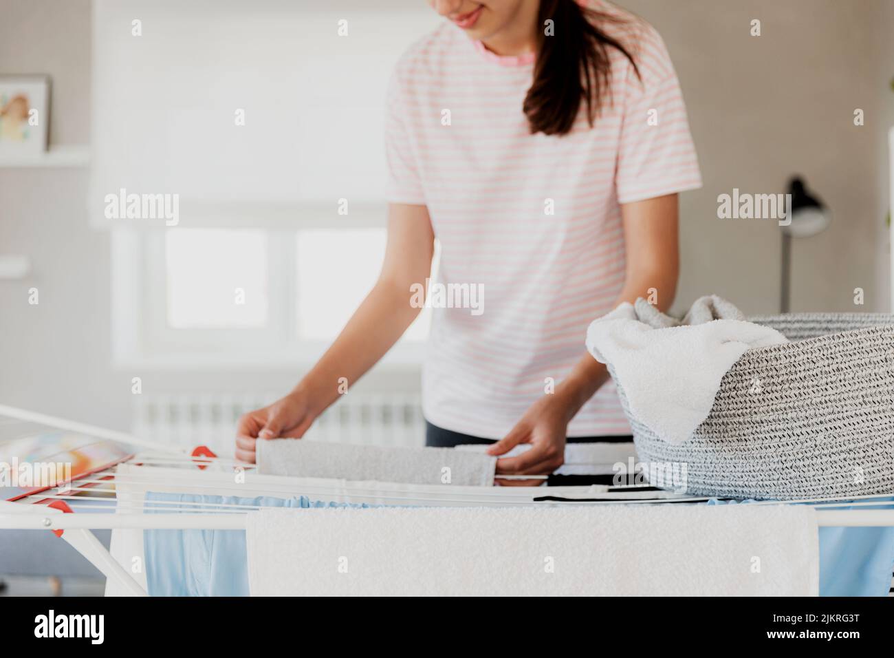 Happy young brunette caucasian woman smiling, hanging clean wet clothes laundry on drying rack at home. Female housewife hands closeup spreading laundry from basket. Girl puts towels after drying them Stock Photo