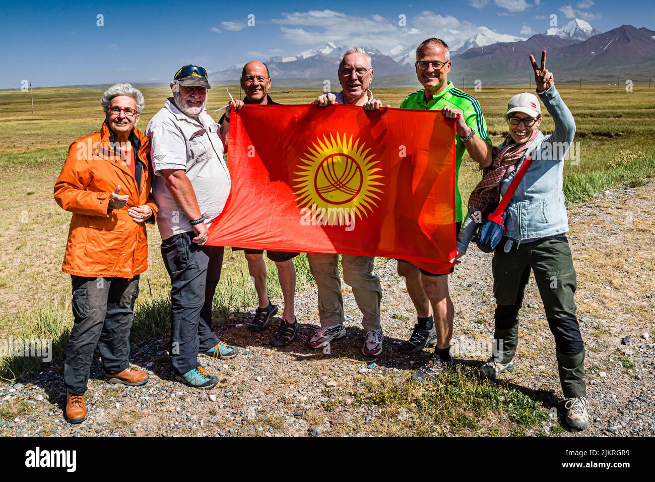 Behind the Kyrgyz flag, three individual travellers and a couple gather around tour organiser Konrad Fobbe (2nd from left), the snow-capped peaks of the Pamir Mountains near Kektyube, Kyrgyzstan, in the background. Stock Photo