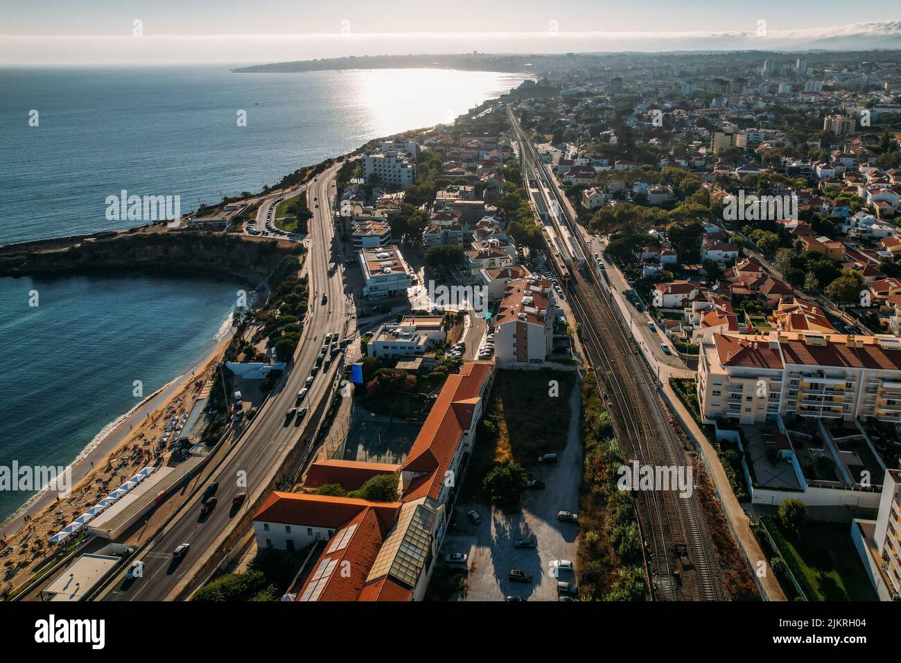 Aerial drone view facing westerwards from Parede, Lisbon Region, Portugal of train at station and coastline during sunset Stock Photo