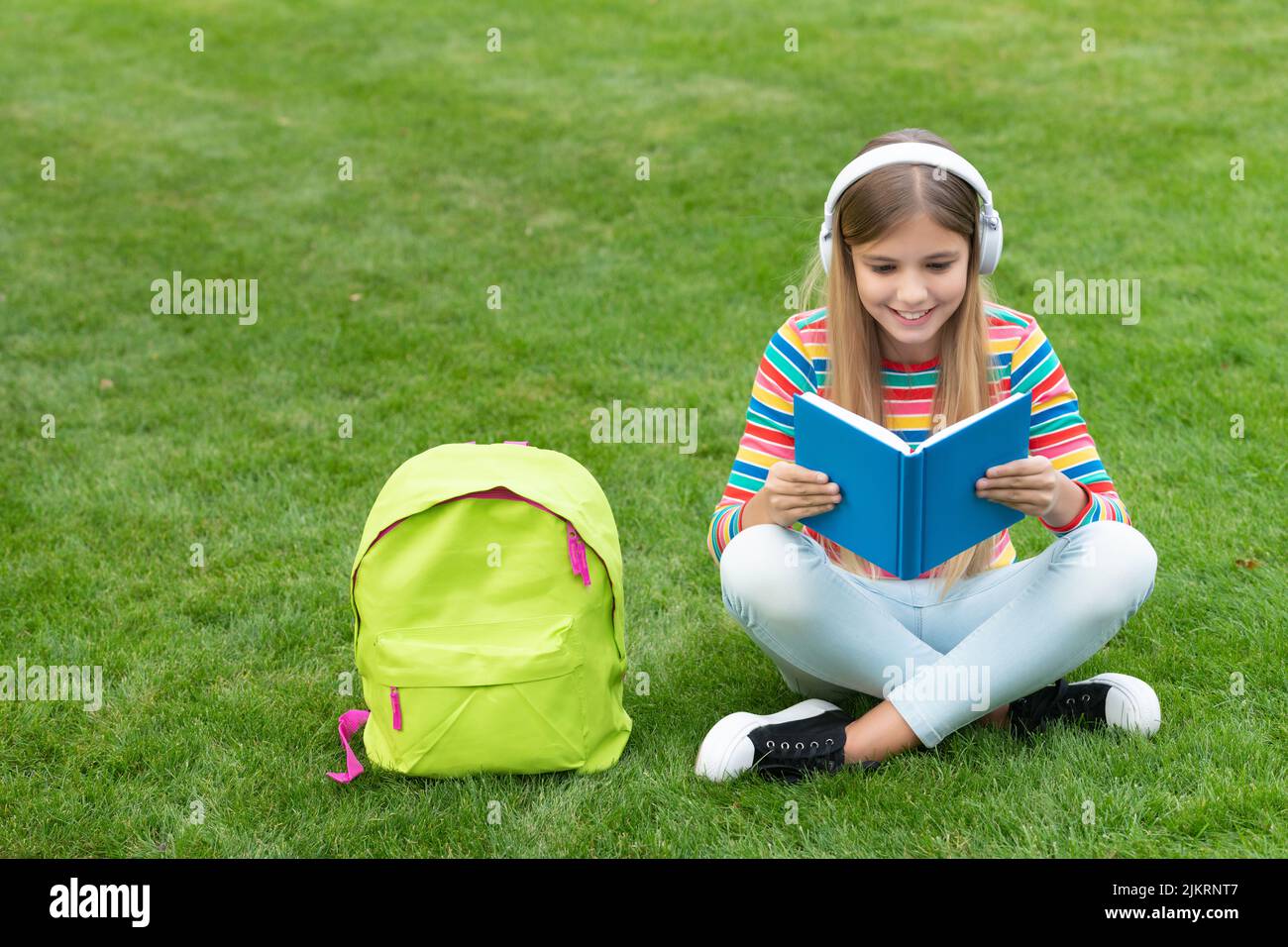 Happy teenage girl reading and listening to audiobook in headphones sitting on grass after school Stock Photo