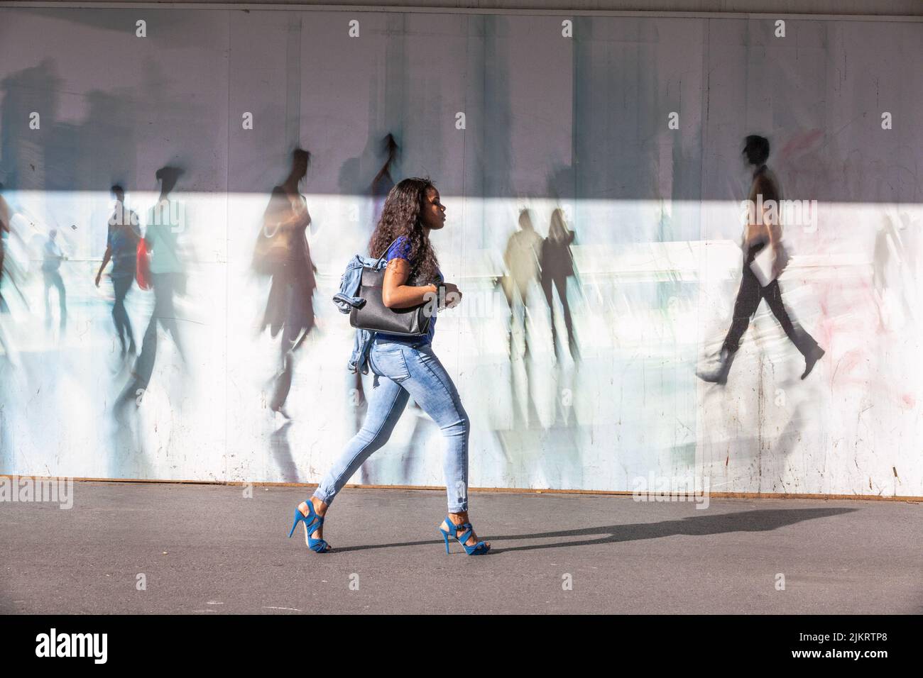 Young woman wearing high heels walking in front of a palisade decorated with figures of passers-by Stock Photo