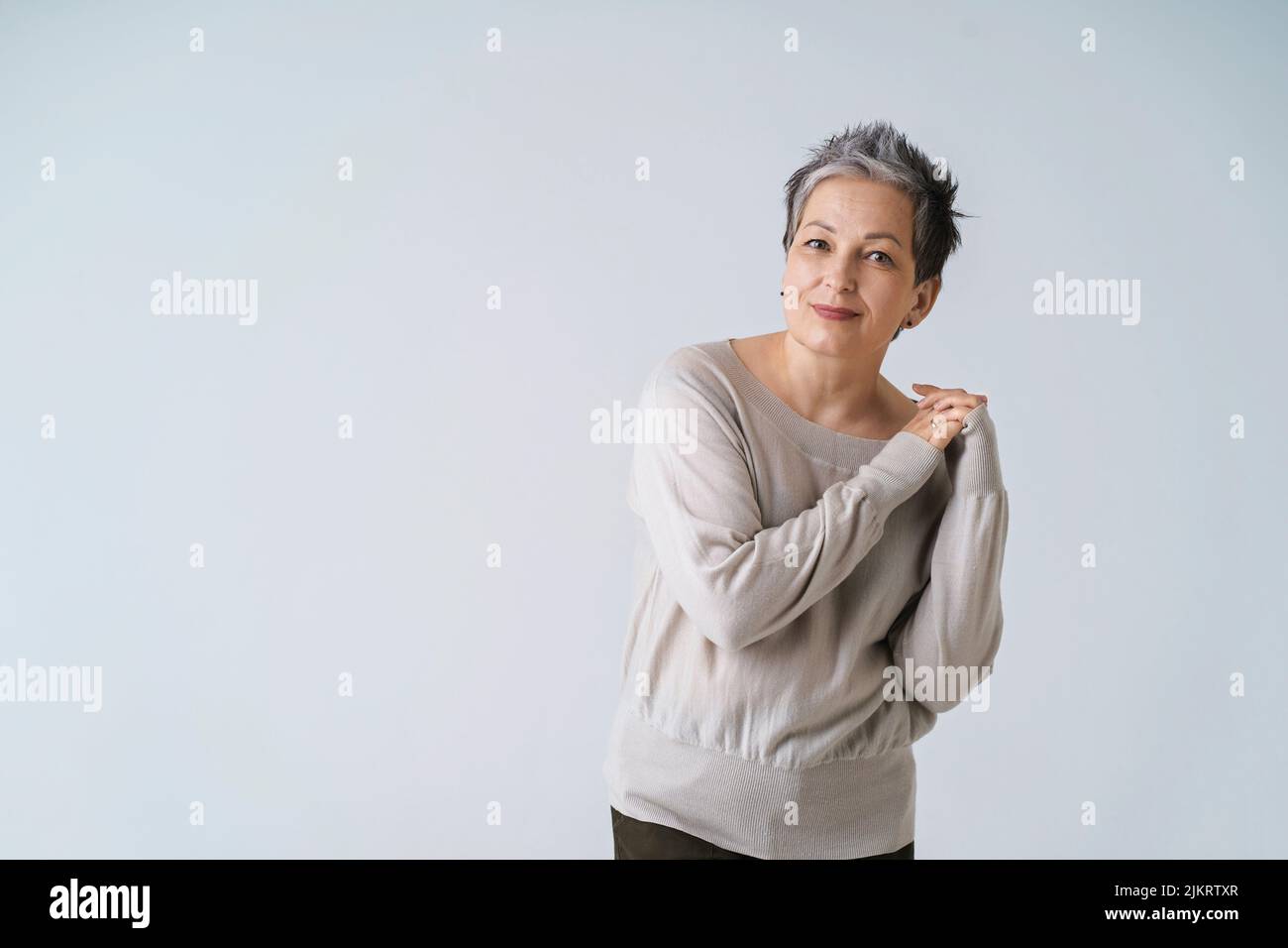 Smiling mature grey short hair woman posing with hands folded at her shoulder looking at camera wearing white blouse, copy space isolated on white background. Healthcare, aged beauty concept.  Stock Photo