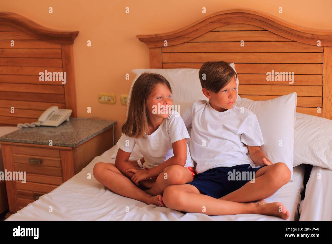 Two children lie on a bed with white sheets in a hotel room on vacation