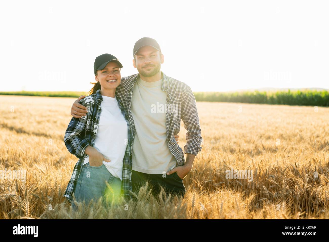 A couple of farmers in plaid shirts and caps stand embracing on agricultural field of wheat at sunset  Stock Photo