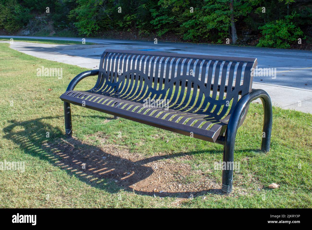 A nice black park bench looks cozy and inviting next to this park side road. Lush green trees in the background provide a peaceful setting. Stock Photo