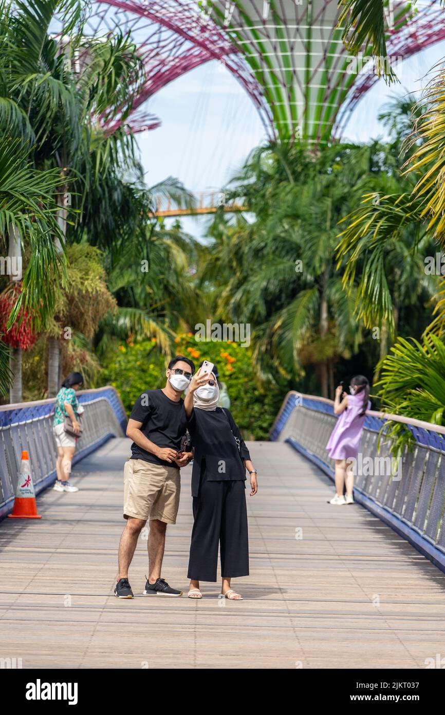 A Malay couple wearing sunshade is taking selfie pictures on the bridge before heading to Gardens by the Bay. Singapore 2022. Stock Photo