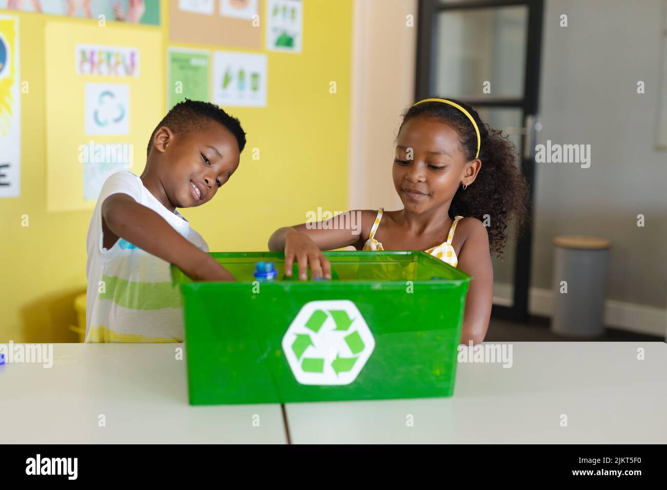 African american elementary students putting bottles in recycling container at desk in school Stock Photo