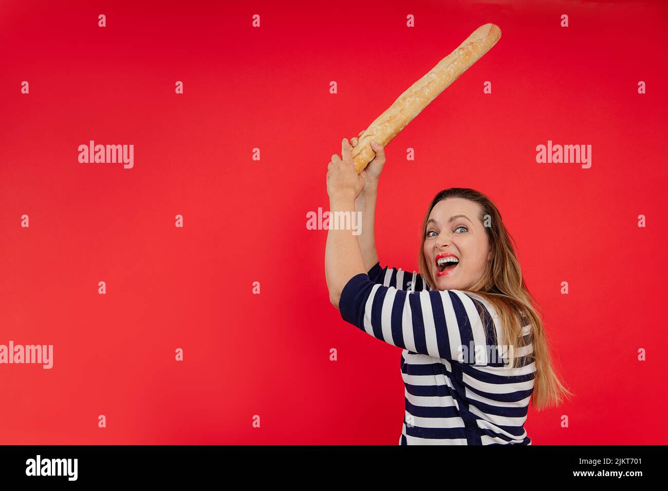 Shouting long haired woman in striped shirt use baguette as sword, cut and breaking discount in red studio. Copy space Stock Photo