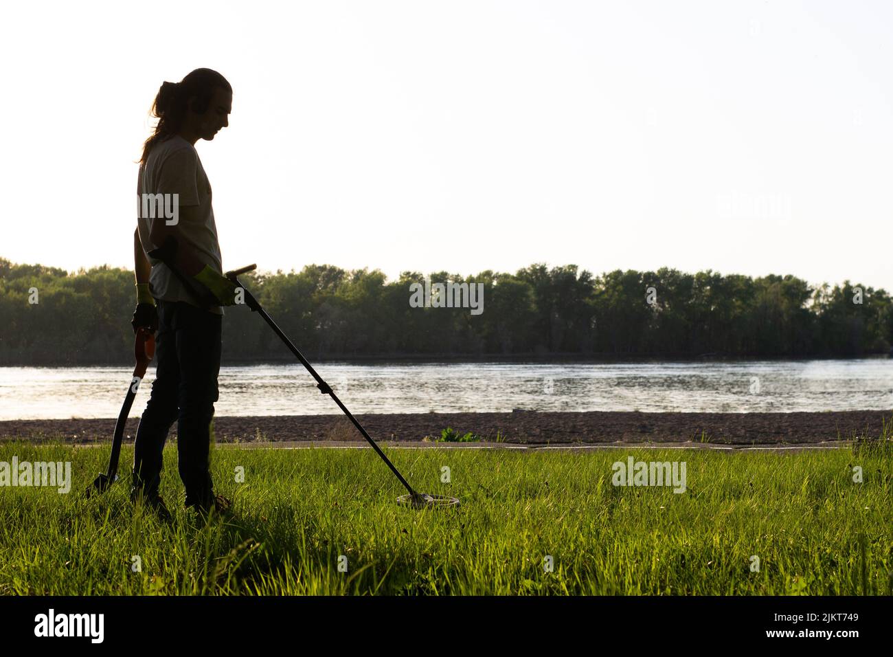 Silhouette of a man with a wireless metal detector on his shoulder and a shovel in his other hand.  On the river bank, trees in the background Stock Photo