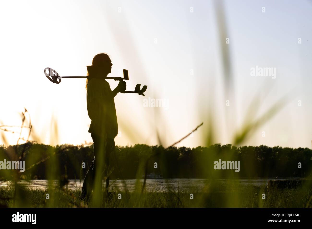 The silhouette of a young man with a wireless metal detector looks ahead.  On the bank of the river, in the foreground the grass is in blur Stock Photo