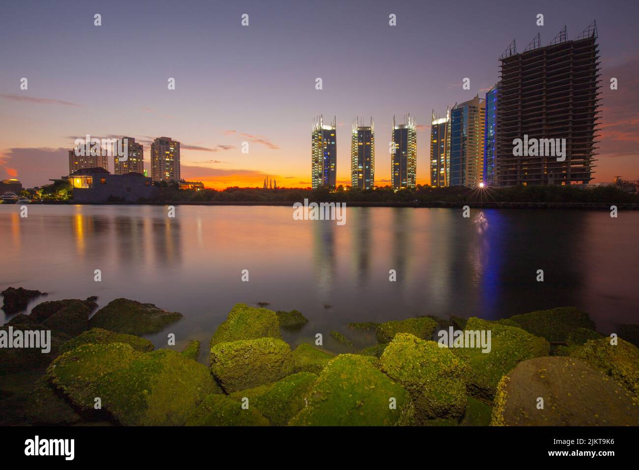 A scenic view of mossy rocks and calm sea against city buildings during sunset Stock Photo