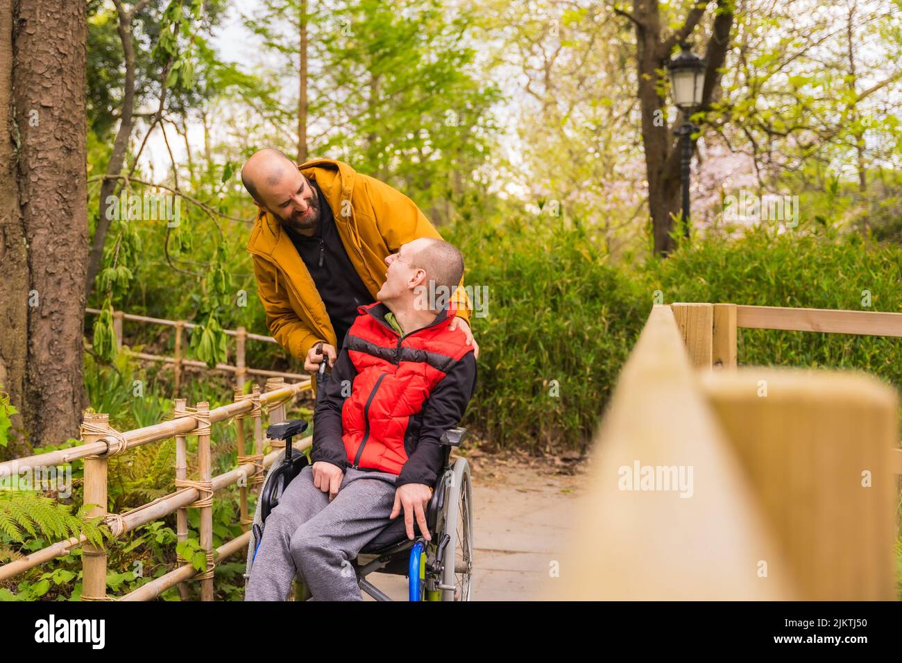 Paralyzed young man in the wheelchair being pushed by a friend in a public city park, looking at each other and smiling Stock Photo